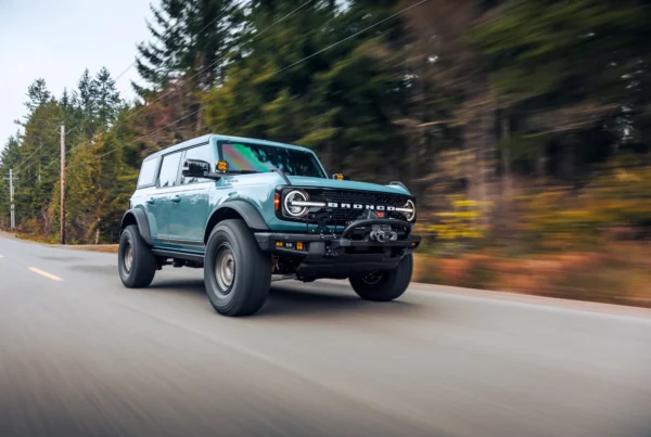 A light blue Ford Bronco SUV with large off-road tires driving along a paved road lined with blurred evergreen and autumn trees.