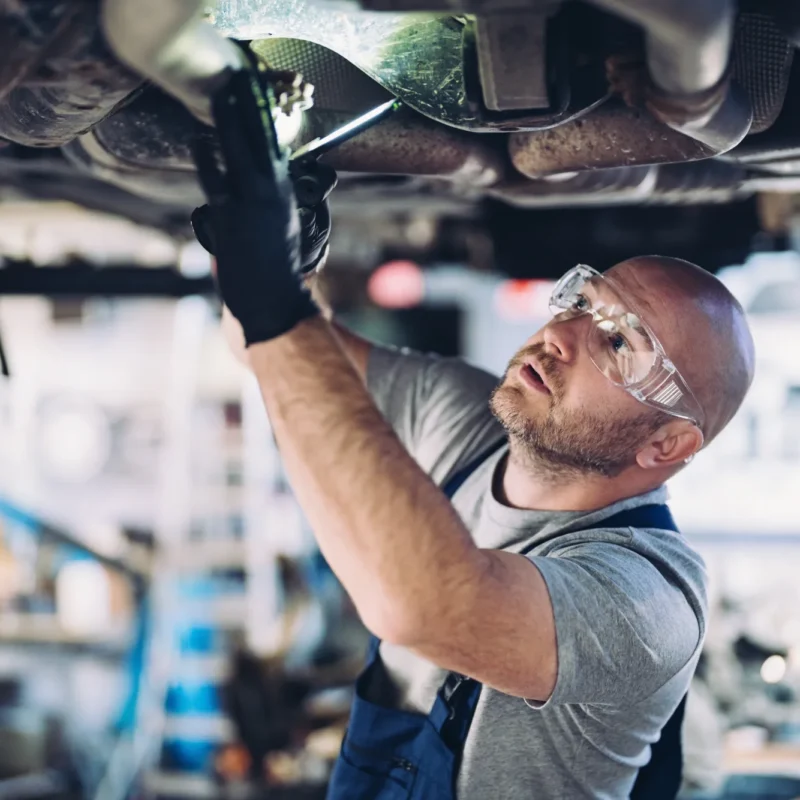 A mechanic in safety glasses and gloves works on the underside of a car raised on a lift in a repair shop.