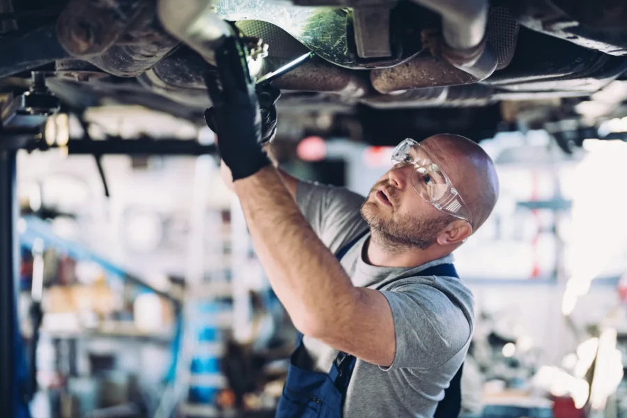 A mechanic in safety glasses and gloves works on the underside of a car raised on a lift in a repair shop.