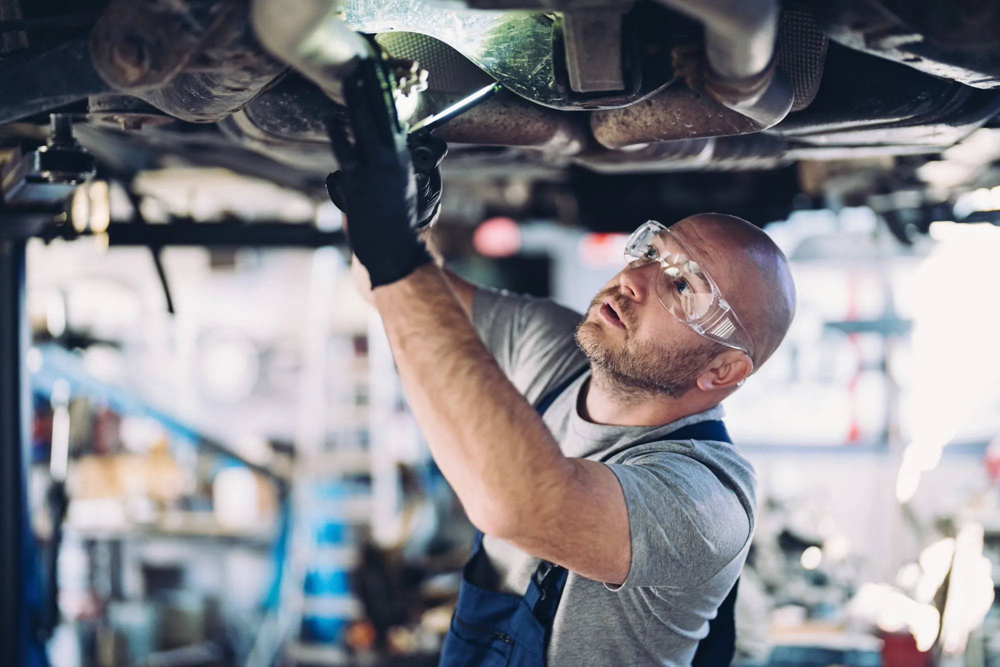 A mechanic in safety glasses and gloves works on the underside of a car raised on a lift in a repair shop.
