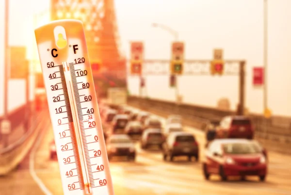 A thermometer showing a high temperature in front of a heat-hazed highway filled with cars.