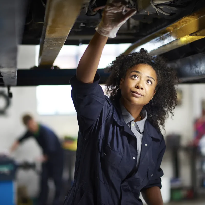 A female mechanic in navy coveralls reaches up to inspect the underside of a car raised on a lift inside an auto repair shop.