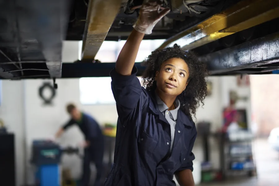 A female mechanic in navy coveralls reaches up to inspect the underside of a car raised on a lift inside an auto repair shop.